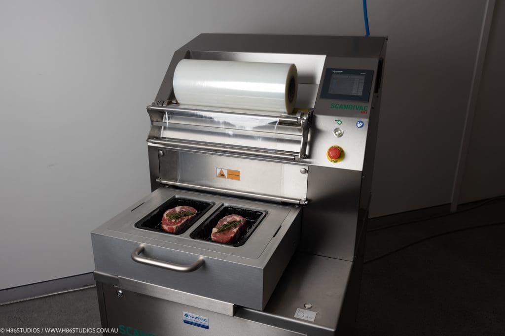 A commercial packaging machine tray sealer preparing to vacuum seal two raw beef steaks in black plastic trays