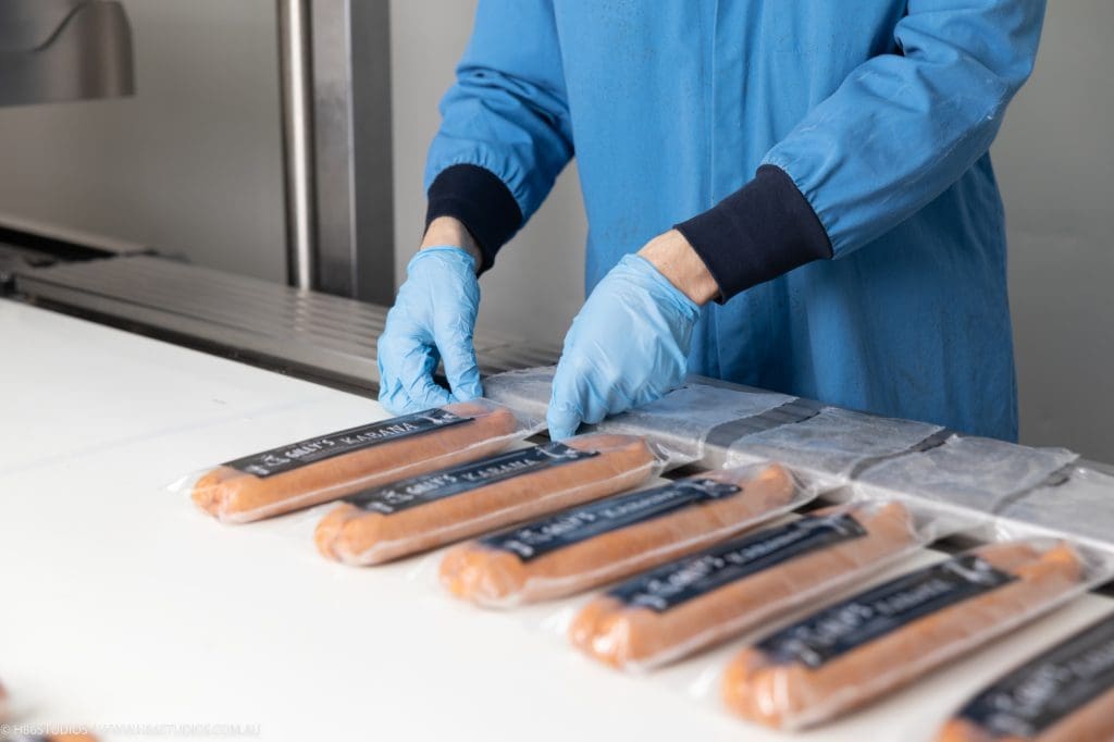 A production worker wearing blue gloves flattens plastic pouches of kabana sausages onto a commercial sealing bar to prevent wrinkles